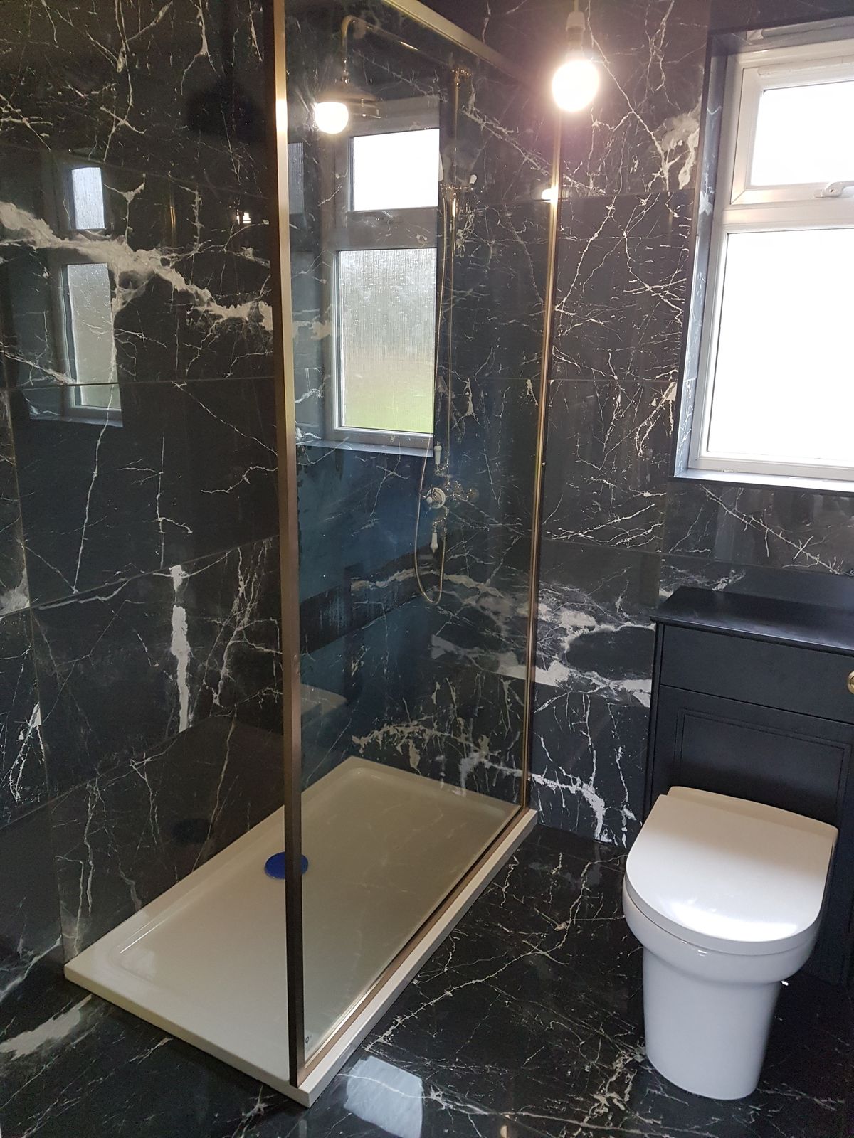 A bathroom tiled floor to ceiling in black marble-effect tiles with a brushed brass-framed shower enclosure