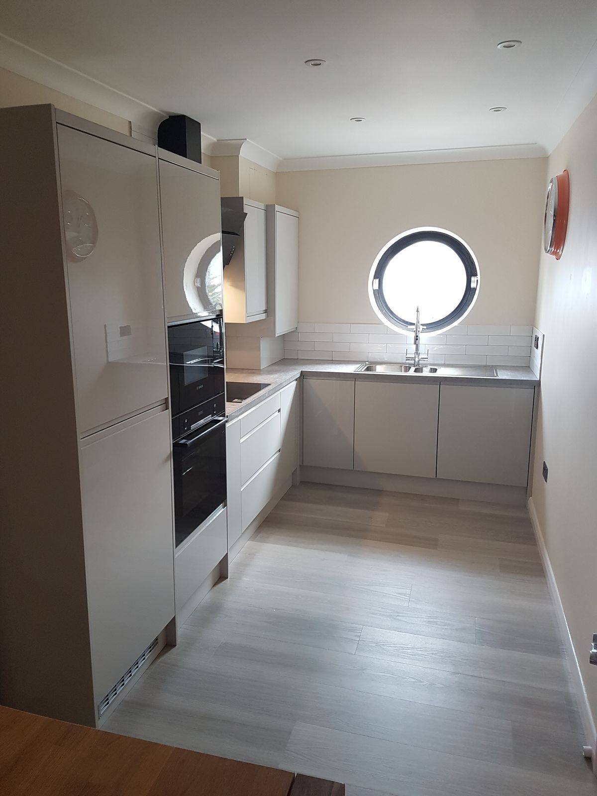 A compact grey gloss kitchen with built-in ovens and a round porthole window
