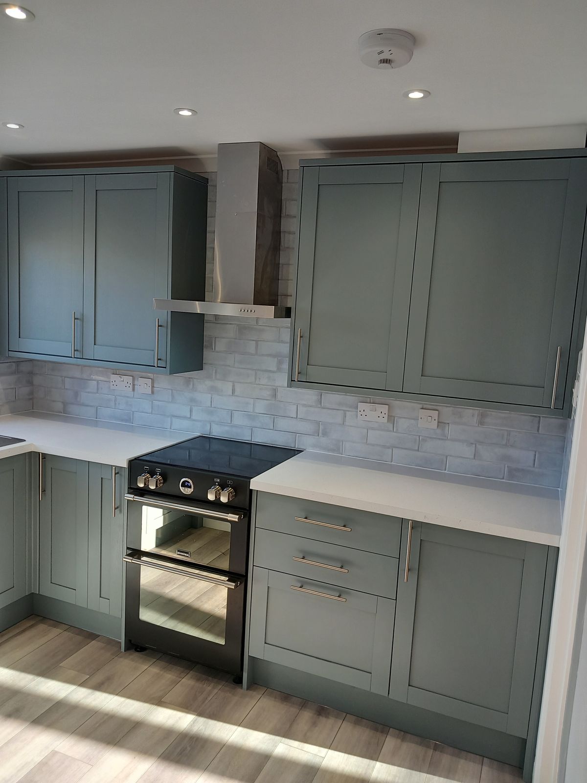 A sage green shaker kitchen with a range cooker, chimney extractor and white tiled splashback